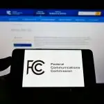 Person holding mobile phone with seal of US agency Federal Communications Commission (FCC) on screen in front of web page. Focus on phone display