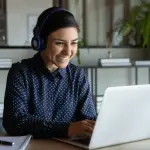 Head shot happy young indian ethnicity female manager wearing wireless headphones, looking at laptop screen, holding pleasant conversation with partners clients online, working remotely at workplace.