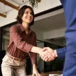 Pay-Per-Click vs. Buying Leads | A woman shakes another man's hand as they both rise up from their seats at an office desk.