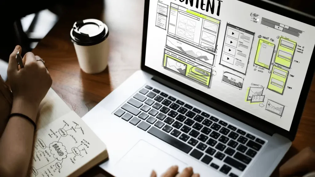 Designer sketching website wireframe layout on laptop screen with brand mind map notebook